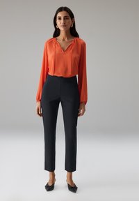 Rood-oranje blouse met lange mouwen, een V-hals en geplooide accenten, gecombineerd met op maat gemaakte zwarte broek en zwarte instapschoenen.