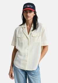 Creme kortærmet skjorte med knaplukning og to frontlommer, parret med lyseblå jeans. Modellen bærer en marineblå og rød kasket med broderet tekst.