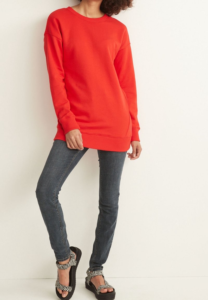 Personne portant un long sweat-shirt rouge, un jean skinny bleu et des sandales noires à plateforme avec des brides imprimées zébrées, debout contre un mur blanc.