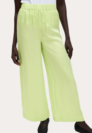 Kvinde iført lysegrønne vidde-ben bukser med elastisk linning og sorte teksturerede slip-on sko mod en ensfarvet baggrund.