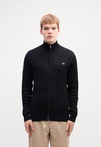 Fekete kötött kardigán, kábelmintás textúrával, magas gallérral, teljes zippel és bordás mandzsettákkal, valamint alján. Márka logó a mellkason.