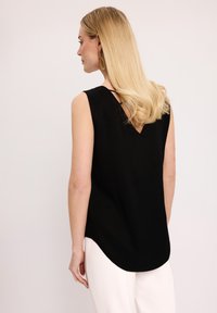 Czarny bezrękawnik z dekoltem w kształcie litery V na plecach, o gładkiej fakturze i zaokrąglonym dolnym wykończeniu, zestawiony z jasnymi spodniami.