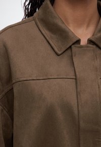 Κοντινό πλάνο ενός ατόμου που φορά καφέ σουέντ μπουφάν με μυτερό γιακά και ορατές λεπτομέρειες ραφών στον ώμο και στο στήθος.