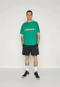 Zöld, rövid ujjú sportpóló, rajta "ADIDAS" felirattal sárga és kék színben, fekete sorttal, fehér zoknival és fekete sportcipővel párosítva.