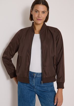 Femme portant une veste bomber marron zippée, une chemise blanche et un jean bleu, debout avec une main sur la hanche devant un fond uni.