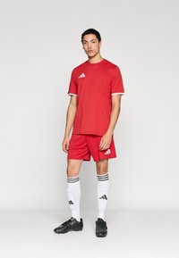 Κόκκινη αθλητική φανέλα με λευκές λεπτομέρειες, συνδυασμένο σορτς, μαύρα ποδοσφαιρικά παπούτσια και λευκές κάλτσες με μαύρες ρίγες και λογότυπο της μάρκας.