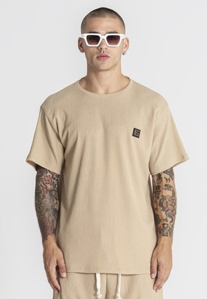 Uomo con tatuaggi che indossa una camicia beige a maniche corte con trama e pantaloni abbinati, occhiali da sole bianchi squadrati, in piedi davanti a uno sfondo semplice.