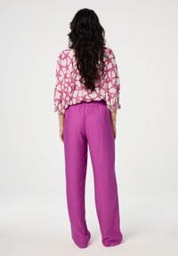 Roze met patroon blouse met pofmouwen, gecombineerd met wijde, felpaarse broek met een elastische tailleband, gefotografeerd van achteren.