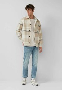 Young man standing, wearing beige plaid jacket, light beige shirt, light blue jeans, and white sneakers against a plain light gray background.