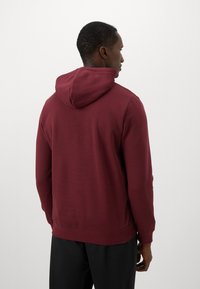 Homme tourné vers l'extérieur portant un hoodie uni bordeaux avec la capuche abaissée et un pantalon noir, sur un fond neutre.