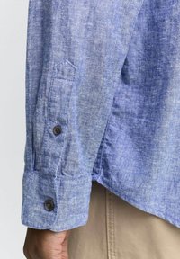 Blauw linnen shirt met een gestructureerde afwerking, voorzien van een knoopmanchet en een afgeronde zoom. De stof heeft een lichte heatherpatroon.