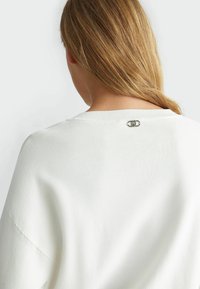 Femme blonde avec les cheveux sur l'épaule, portant un pull en maille blanc présentant un petit détail de logo en argent au niveau de l'encolure arrière.