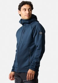 Man wearing a navy blue zip-up hoodie with a hood, standing and looking to his left against a white background.