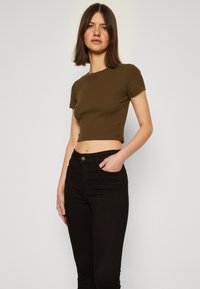 Oliivinvihreä lyhythihainen crop-top, joka on yhdistetty korkeavyötäröisiin mustiin farkkuihin. Yläosassa on tiukka, uritettu pinta.