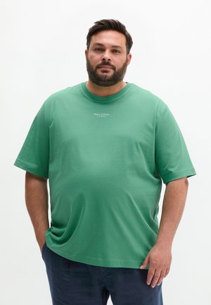 Mężczyzna z brodą w zielonej koszulce Marc O'Polo i ciemnych spodniach stojący na jednolitym białym tle.