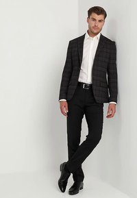 Blazer a quadri in sfumature di grigio con accenti marroni, camicia bianca, pantaloni neri e scarpe eleganti nere, in piedi contro un muro bianco.