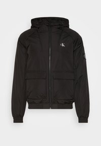 Svart puffjacka med huva, med dragkedjestängning, två framfickor, elastiska ärmslut och en logotyp på bröstet och ärmen.