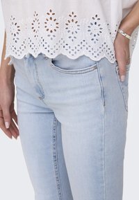 Egy világos kék denim farmernadrág, slim fit fazonban, fehér felsővel párosítva, amelynek szegélyén hímzett lyukacsos részletek találhatóak.