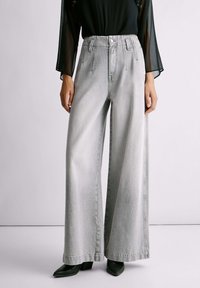 Kobieta w jasnoszarych, wysokich szerokich dżinsach z przednimi szwami, połączonych z czarną, przezroczystą bluzką z długim rękawem oraz czarnymi, spiczastymi butami.