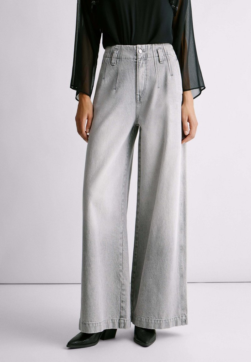 Kobieta w jasnoszarych, wysokich szerokich dżinsach z przednimi szwami, połączonych z czarną, przezroczystą bluzką z długim rękawem oraz czarnymi, spiczastymi butami.