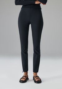 Schwarze, figurbetonte Hose mit glattem Stofffinish, hoher Taille und knöchellangem Schnitt; kombiniert mit schwarzen Riemchensandalen.