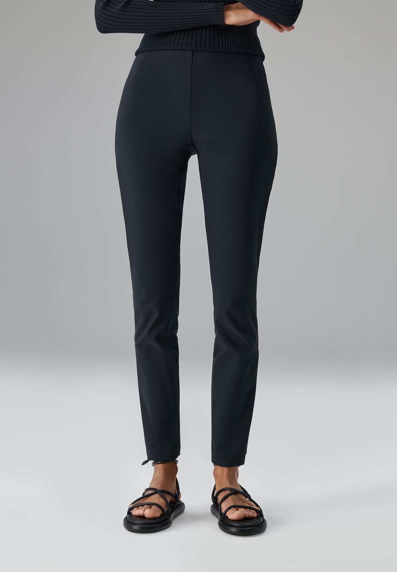 Schwarze, figurbetonte Hose mit glattem Stofffinish, hoher Taille und knöchellangem Schnitt; kombiniert mit schwarzen Riemchensandalen.