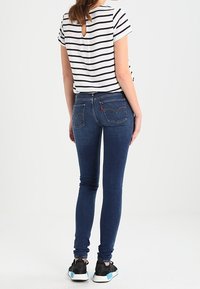 Una persona está de espaldas a la cámara, vistiendo una camisa a rayas y jeans ajustados azules, combinados con zapatillas negras.