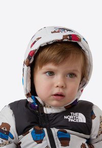 Jachetă matlasată cu model de urs alb, cu fermoar negru în față și glugă. Logo-ul The North Face este afișat pe piept.