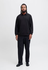 Sort sweatshirt med krave, afslappet pasform, struktureret stof, lagdelt over en rød skjorte; kombineret med løse sorte jeans og sorte sko.