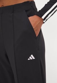 Zwarte sportbroek met een gladde textuur, voorzien van een wit Adidas-logo aan de linkerkant en zakken aan beide zijden.