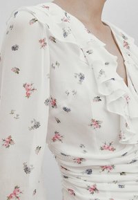 Blouse blanche avec imprimé floral comportant de petites fleurs roses et bleues. Encolure froncée et corps ajusté avec des fronces. Tissu doux et léger.