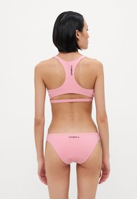 Růžový bikinový set vyrobený z hladkého materiálu. Vrchní část má design s průřezem na zádech a výřezy; spodní část má nízký pas a na zadní straně logo.