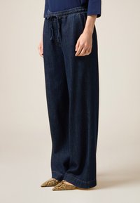 Pantalones de mezclilla de pierna ancha en azul oscuro, con una cinturilla elástica y cordón. La textura es suave y los puños están rematados.
