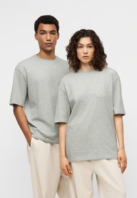 Άνδρας και γυναίκα φορούν ταιριαστά γκρι φαρδιά μπλουζάκια και ανοιχτό μπεζ παντελόνια, στέκονται κοντά ο ένας στον άλλον μπροστά σε απλό λευκό φόντο.