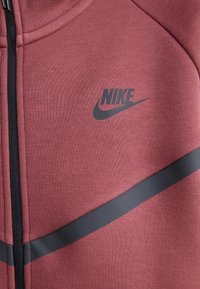 Casaco Nike com fecho éclair em vermelho suave, com um acabamento texturizado. Apresenta um logo cinza escuro e detalhes de riscas horizontais.