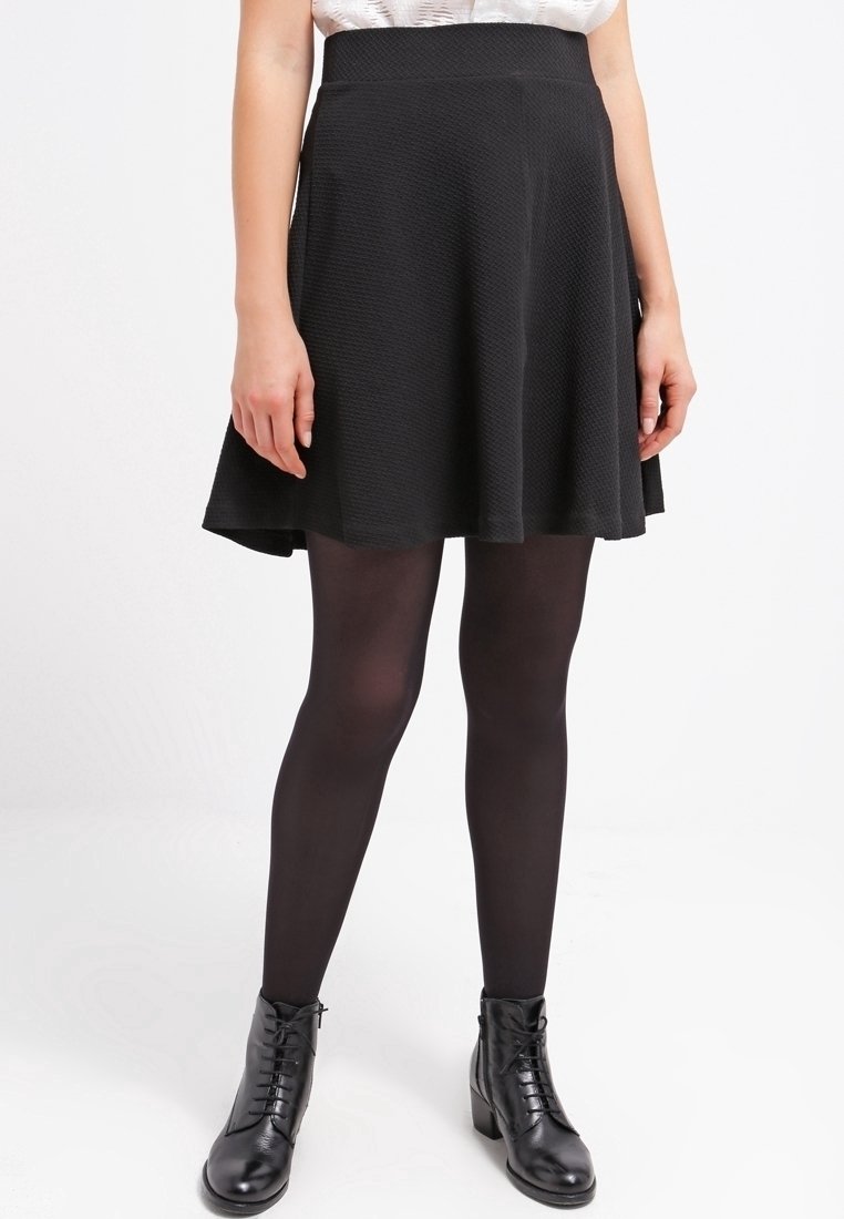 Femme portant une jupe évasée texturée noire, des collants noirs et des bottines noires à lacets, debout devant un fond clair uni.