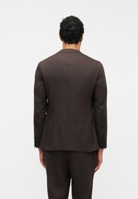 Άνδρας με κοντά σγουρά μαλλιά, ντυμένος με εφαρμοστό σκούρο καφέ κοστούμι, στέκεται γυρισμένος με την πλάτη σε λευκό απλό φόντο.