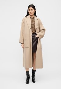 Γυναίκα που φοράει μπεζ μακρύ παλτό, τοπ με τύπωμα λεοπάρδαλης, σκουρόχρωμη καφέ δερμάτινη φούστα και μαύρα μποτάκια στον αστράγαλο, στέκεται μπροστά σε λευκό φόντο.