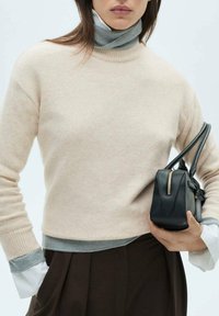 Maglione beige chiaro realizzato in materiale morbido, con dettagli grigi sui polsini e sull'orlo, abbinato a una piccola borsa nera con zip dorata.