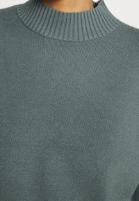Turkusowy sweter z dzianiny z prążkowanym, wysokim dekoltem, charakteryzujący się teksturowanym materiałem i luźnym krojem. Brak widocznych wzorów lub akcentów.