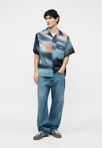 Joven con cabello rizado y oscuro, vistiendo una camisa de botones de manga corta con estampado, jeans azules holgados y zapatillas verdes, de pie con una mano en el bolsillo.