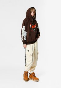 Joven vestido con una sudadera gráfica marrón, pantalones cargo crema con bolsillos y correas, y botas beige con cordones, de pie contra un fondo blanco.