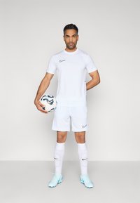 Λευκή αθλητική φανέλα και σορτς Nike, κρατώντας μια ποδοσφαιρική μπάλα με σχέδιο. Περιλαμβάνει μαύρα λογότυπα, κάλτσες μέχρι το γόνατο και γαλάζια παπούτσια ποδοσφαίρου.