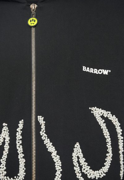 Μαύρο ένδυμα με φερμουάρ και ασημί φερμουάρ; η λαβή του φερμουάρ έχει πράσινο χαμογελαστό πρόσωπο με δόντια; λευκό κείμενο "BARROW" και σχέδιο μαργαρίτας φλόγας.