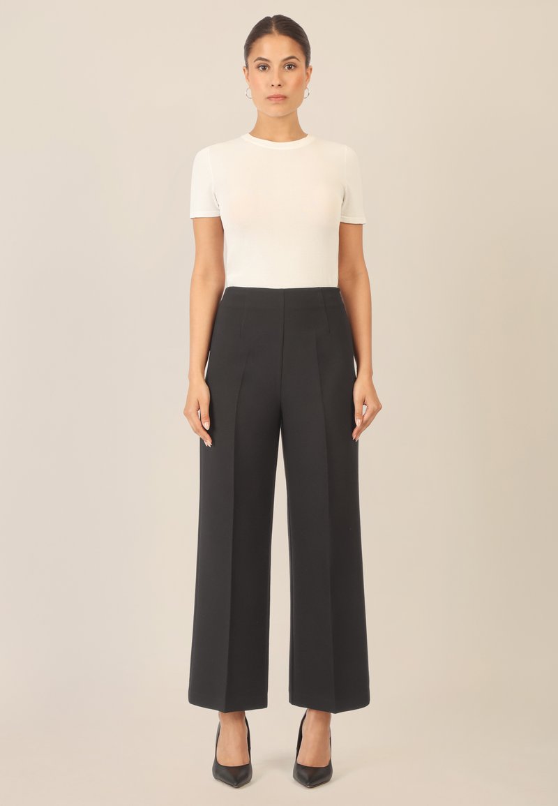Pantalons plissés noirs taille haute avec jambes larges, associés à un haut ajusté blanc à manches courtes et des escarpins noirs à bout pointu.