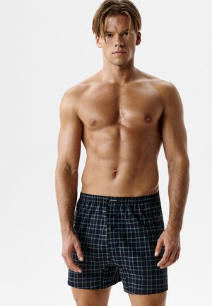 Młody muskularny mężczyzna w czarnych, kraciastych bokserkach stoi na tle jednolitego, białego tła.