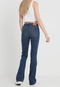 Žena s dlouhými blond vlasy, oblečená v bílém bezramenném topu a modrých bootcut džínách, stojící před jednobarevným pozadím.