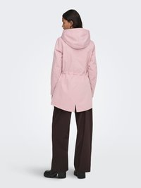 Femme portant une veste à capuche rose clair avec une taille cintrée, un pantalon large et sombre, ainsi que des bottes noires épaisses, dos tourné à l'appareil photo.