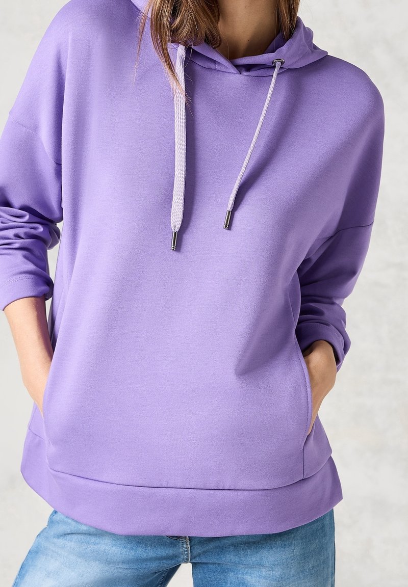 Femme portant un sweat à capuche lilas avec cordons et poches avant, associé à un jean bleu clair, mains dans les poches.