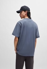 Tricou gri din bumbac cu mâneci scurte, croială lejeră, decolteu rotund și design simplu. Purta cu pantaloni închiși la culoare și o șapcă neagră.
